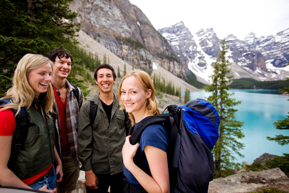 Vier lächelnde Kollegen, ausgestattet mit Rucksäcken, stehen vor einem kristallklaren Bergsee mit malerischen, schneebedeckten Gipfeln im Hintergrund. Sie scheinen eine Pause auf ihrer Wanderung zu genießen und strahlen Freude und Zufriedenheit aus.