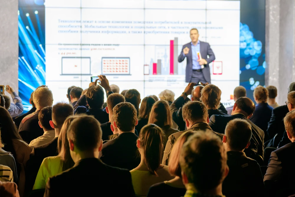 Eine Menschenmenge, aus dem Blickwinkel der Zuhörer fotografiert, lauscht aufmerksam einem Redner, der vor einem großen Bildschirm mit grafischen Darstellungen steht. Die Szene spielt sich in einem beleuchteten Konferenzsaal ab.