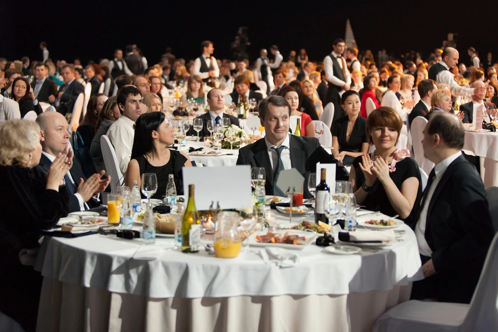 Das Bild zeigt Gäste an gedeckten Tischen bei einer formellen Veranstaltung oder Gala. Die Teilnehmer scheinen in Gespräche vertieft zu sein oder dem Programm zu folgen, während sie umgeben von Geschirr und Getränken sitzen.