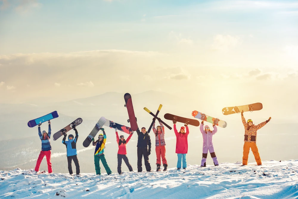 Eine Gruppe fröhlicher Snowboarder steht auf einem schneebedeckten Berg und hält ihre Boards triumphierend in die Luft, mit einer malerischen Winterlandschaft und strahlendem Sonnenlicht im Hintergrund.