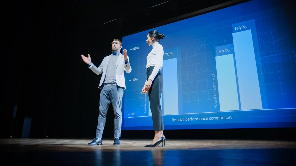 Das Bild zeigt zwei Personen, die eine Präsentation auf einer Bühne halten, wobei ein großer Bildschirm hinter ihnen eine Balkendiagramm-Grafik anzeigt. Sie scheinen das Publikum durch ihre Präsentation zu führen.