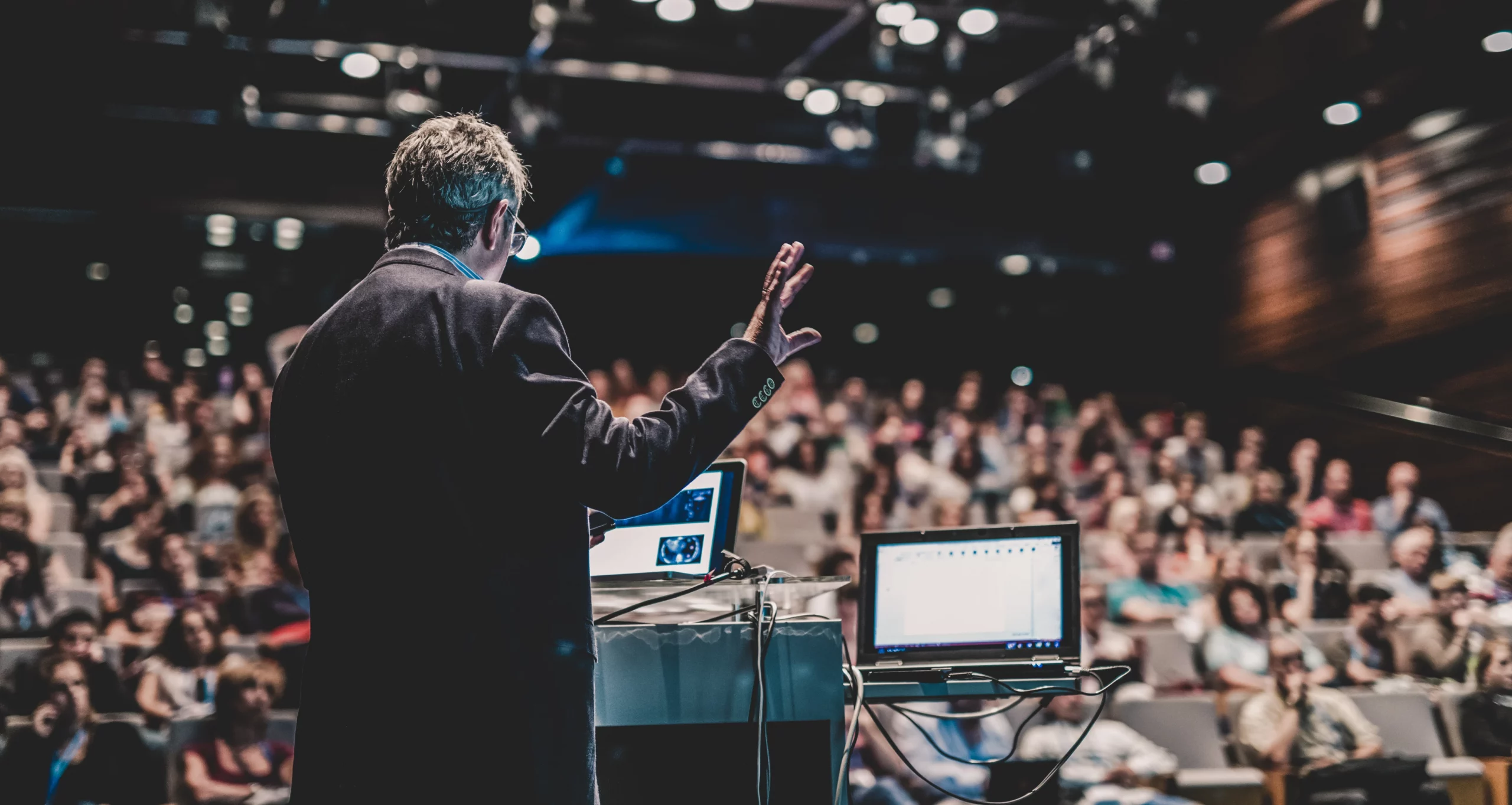 Das Bild zeigt einen Sprecher, der vor einem Laptop auf einem Podium steht und eine Präsentation hält. Er spricht zu einem Publikum in einem großen Hörsaal, das unscharf im Hintergrund zu sehen ist.