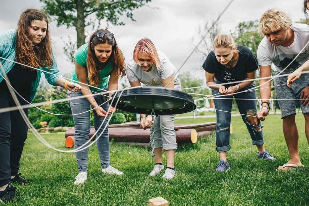 Das Bild zeigt eine Gruppe junger Menschen, die im Freien an einer Teambuilding-Übung teilnehmen. Sie konzentrieren sich darauf, gemeinsam eine Aufgabe zu lösen, bei der sie scheinbar einen Gegenstand mit Seilen manipulieren.