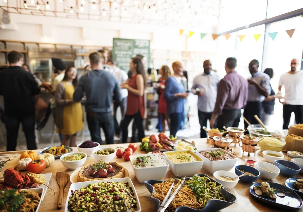 Das Bild zeigt ein Buffet mit einer Vielzahl von Speisen im Vordergrund und eine Gruppe von Menschen, die im Hintergrund in einem hellen und modernen Raum gesellig beisammenstehen.