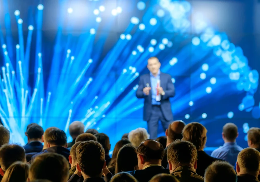 Ein Redner steht auf einer Bühne mit dynamischer blauer Beleuchtung und digitalen Effekten, während er vor einem Publikum aus Geschäftsleuten spricht, die ihm aus der Perspektive der Zuhörer zugewandt sind.